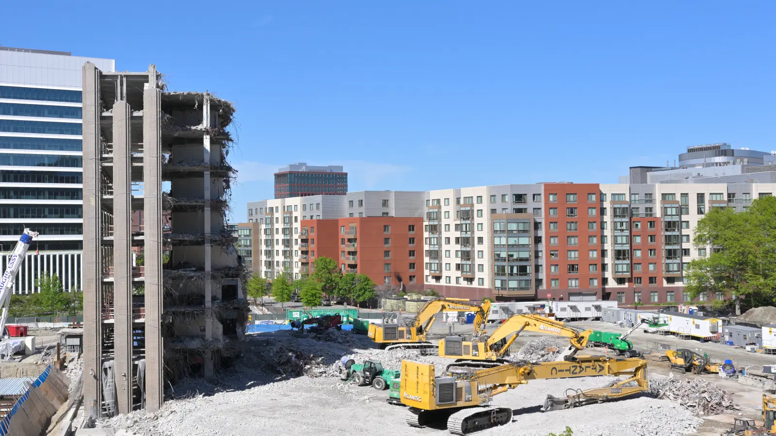 A concrete building being torn down by excavators, with apartment buildings standing behind it